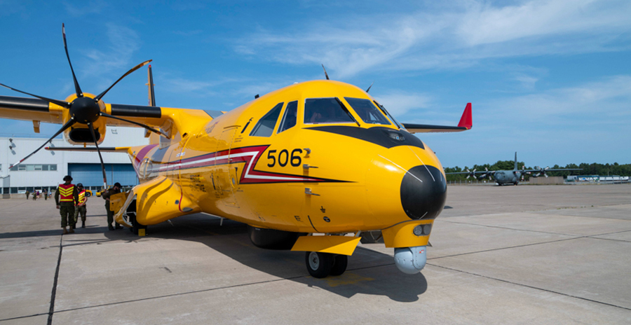 A search and rescue aircraft is parked on an airfield, with a few uniformed people standing nearby. Other planes are in the background.