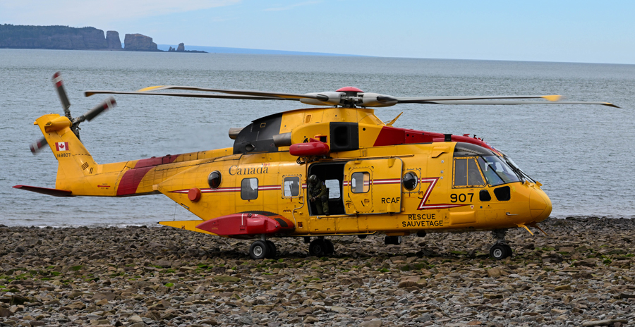 A yellow and red search and rescue helicopter is parked on a rocky shoreline with the ocean and distant cliffs in the background.