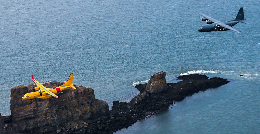 A yellow and red aircraft flies low over rocky outcrops in the ocean, while a gray military transport plane approaches from the right.