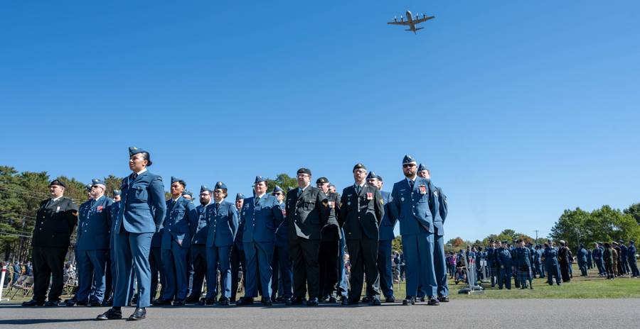 A group of uniformed military personnel stands in formation outdoors under a clear blue sky as a large aircraft flies overhead.