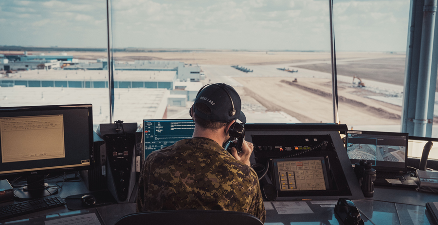 An RCAF member sits at a bank of computers in a tower overlooking an airfield.