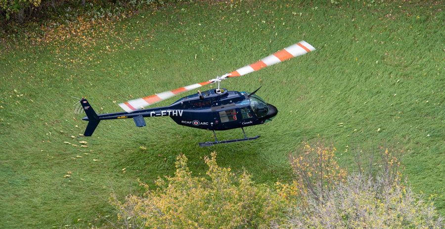 A blue helicopter with red and white rotor blades lands in a grass field surrounded by trees.