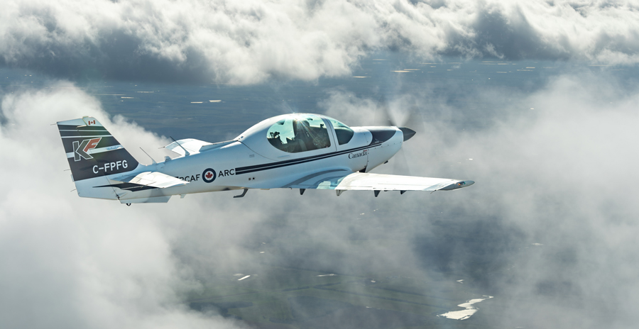 A small white plane in the sky against a backdrop or white/grey clouds with fields and lakes below.