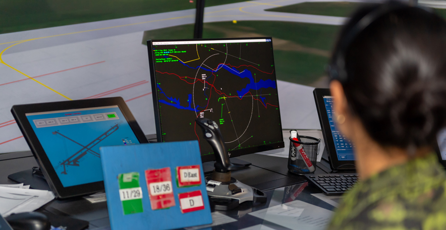 A person in green camouflage is seen looking over computer screens on a counter with a computer-generated airfield behind.