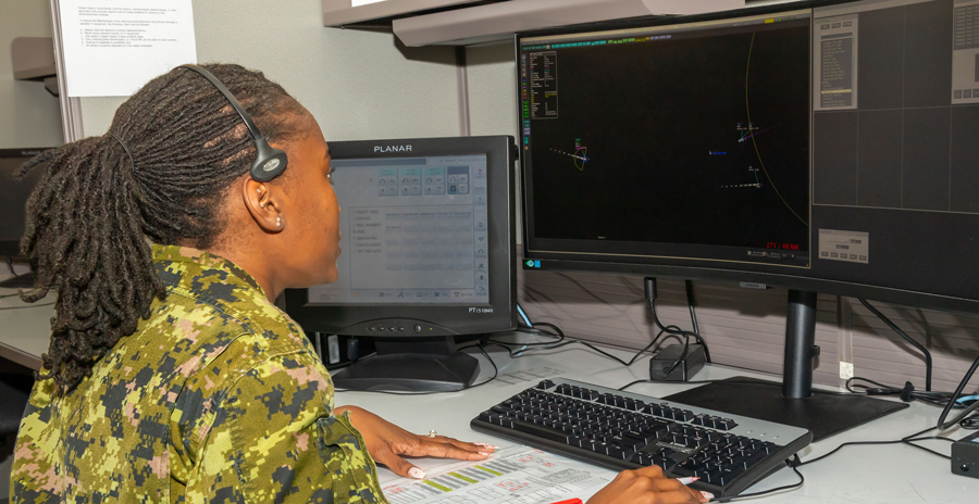 A person in green camouflage wearing a headset looks at computer screens behind a desk.