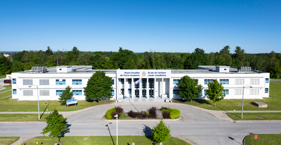 A large grey building with a blue flag and road out front is seen in front of a wooded area.