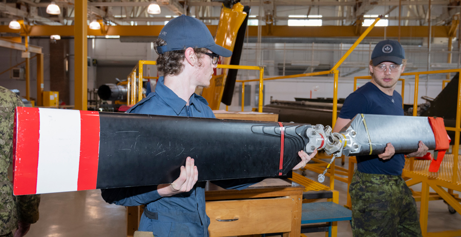 Two people dressed in military clothing carry a tail rotor that is black with red and white tips inside a hangar.