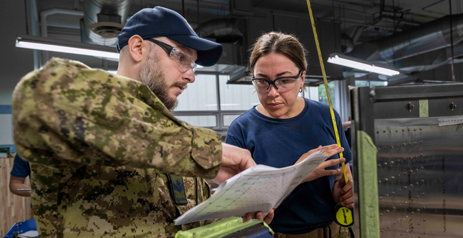 Two military members look at instructions on a paper. To the right, a sheet of silver metal is held on a jig.