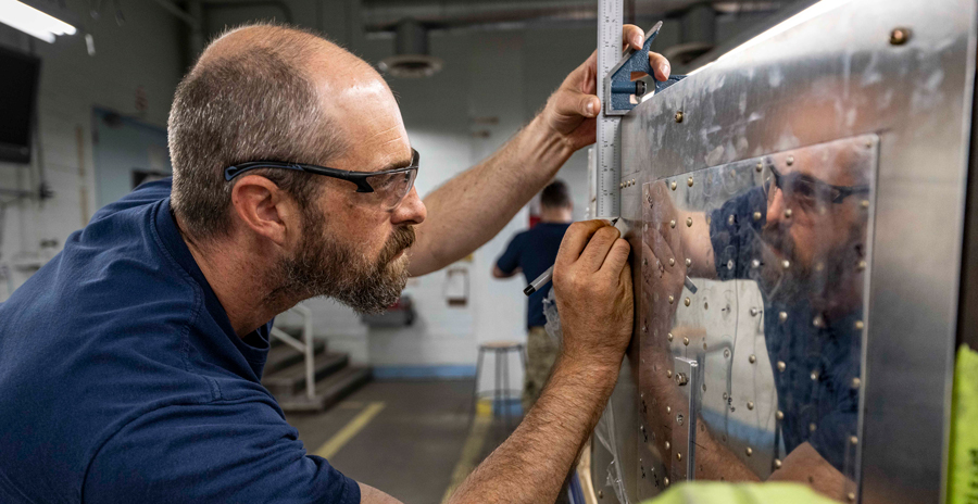 A person in a blue t-shirt uses a pen to mark a piece of silver metal held on a table in a jig.