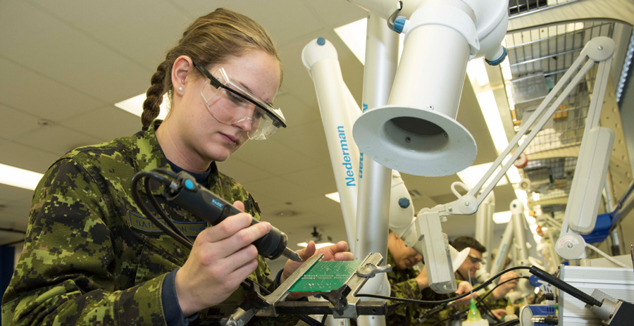 A person in green camouflage uniform and safety glasses stands at a desk with a green plate in a jig. They hold a black soldering iron.