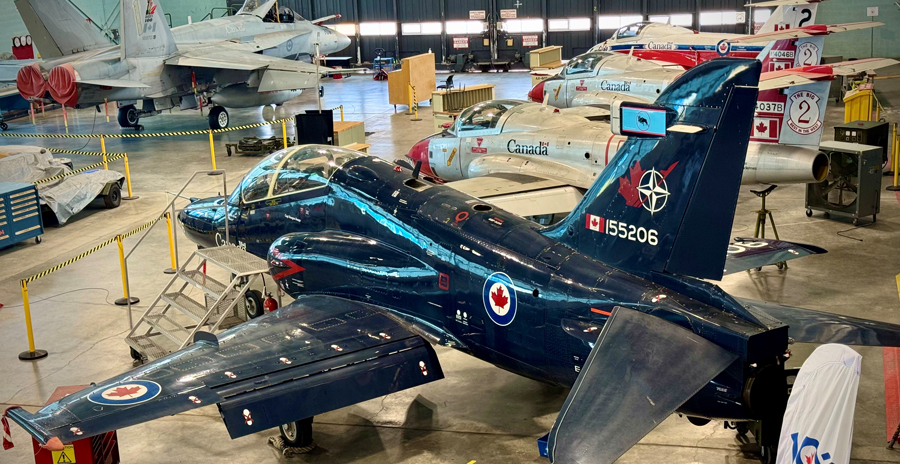 A hangar containing several aircraft is pictured with a blue, two-seat aircraft in the foreground.