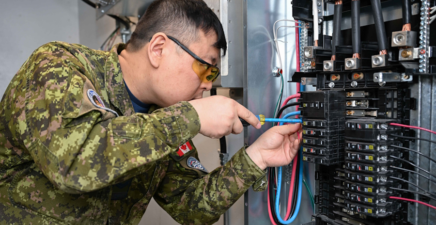 A military member in a camouflage uniform works on wiring inside an electrical generator panel using a screwdriver.