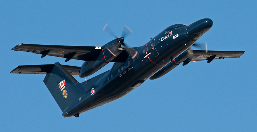 A high-winged aircraft painted dark blue with “Canada” and the number “803” visible on the fuselage flies against a clear blue sky.