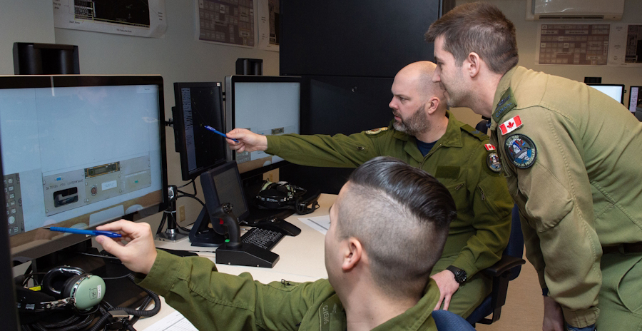 Three military members in green flight suits gather around computer screens, while the two seated members point to information displayed.