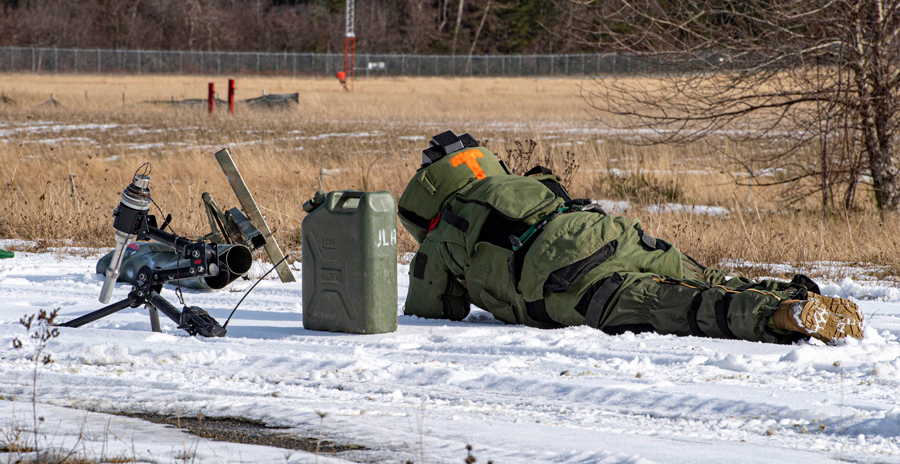 Bomb disposal technician in protective suit lying on snow near a robot and fuel can in an open field.