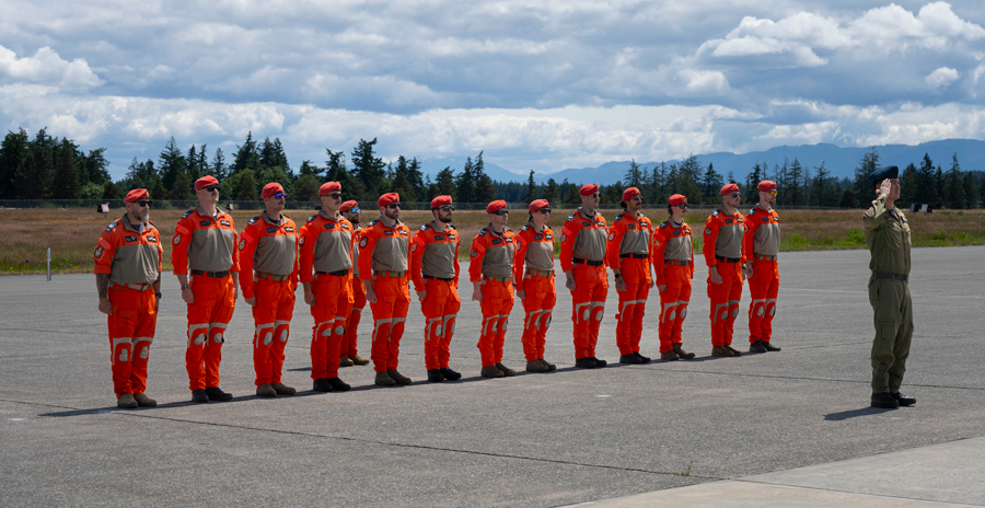 Thirteen Search and Rescue Technicians in orange uniforms stand in formation on a tarmac during a graduation ceremony.