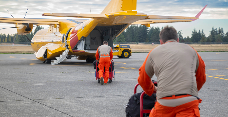 Search and Rescue Technicians push gear carts toward a yellow CC-295 Kingfisher aircraft on a tarmac during SAREX 2025.