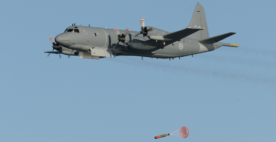 Grey CP-140 Aurora aircraft in flight releasing a training torpedo with parachute against a clear blue sky.