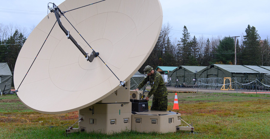Military personnel work on a large deployable satellite system outdoors, with equipment cases and a dish antenna visible on site.