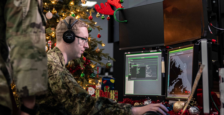 Military personnel work at computer stations decorated for Christmas with NORAD Santa Tracker displayed on a large screen.