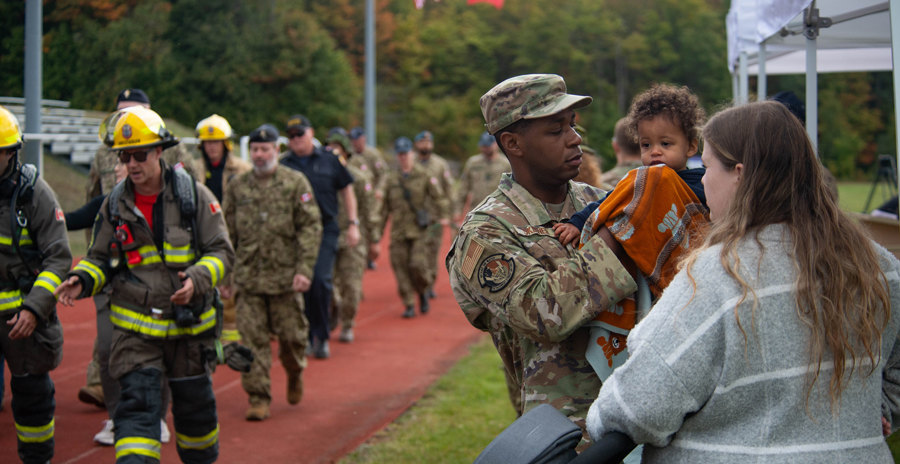 Emergency responders and military members walk along a track while a service member interacts with a family nearby.