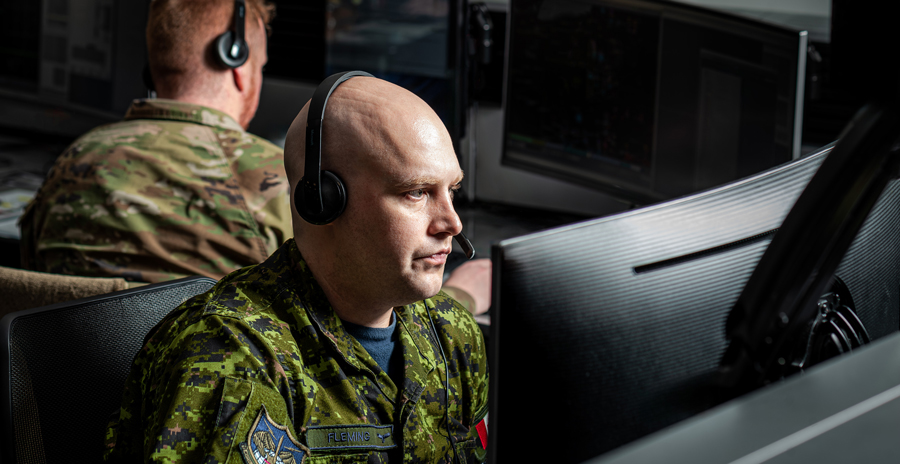 Military personnel in camouflage uniforms work at computer stations and monitor multiple screens.