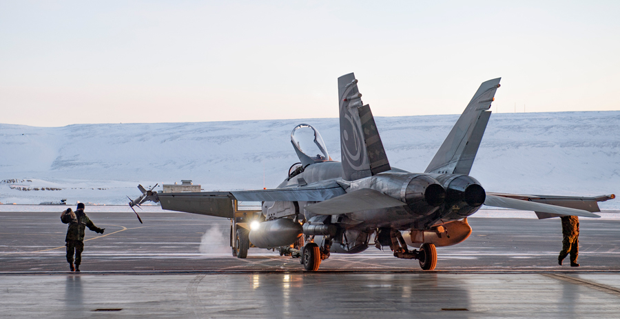 Military jet on snowy tarmac with open canopy, crew performing checks amid steam in cold, remote environment.