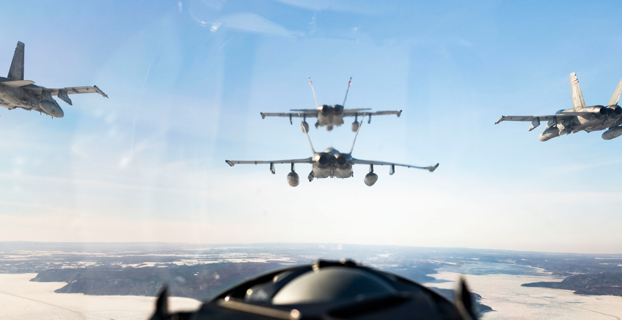Four fighter jets flying in formation over a snowy landscape, viewed from inside another aircraft cockpit.