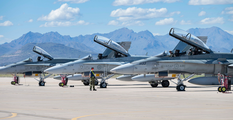 Three fighter jets parked on a runway with open canopies, mountains in the background and a person standing nearby.