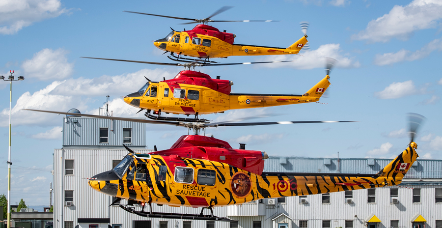 Three yellow and red rescue helicopters hover in formation near a hangar on a clear day with blue skies.