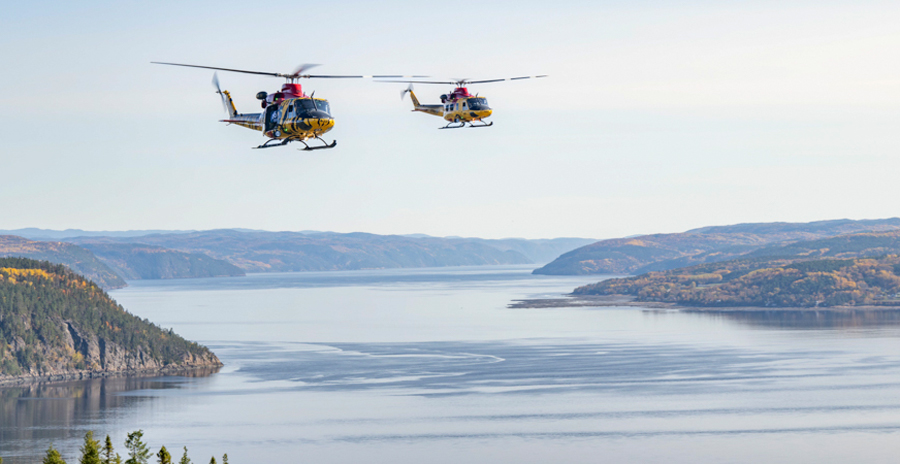 Two yellow rescue helicopters flying over a wide river valley with forested hills and calm water below.