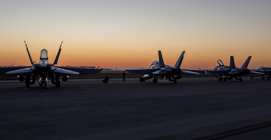 Five fighter jets parked on a runway at sunset, silhouetted against an orange sky.