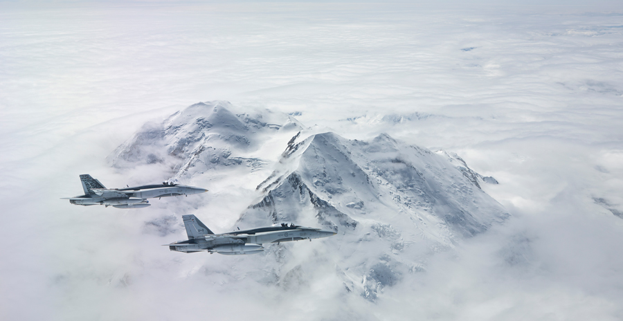 Two fighter jets flying over snow-covered mountains surrounded by clouds under a clear sky.