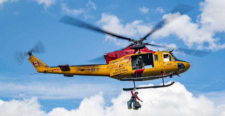 Yellow and red rescue helicopter performing a hoist operation with a person suspended below against a cloudy sky.