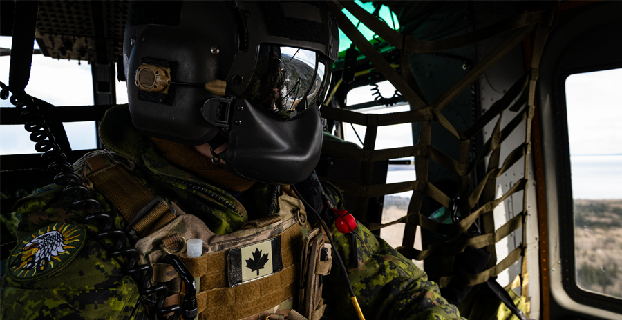 A member of the Canadian Armed Forces in a camouflage uniform and tactical gear sits inside a helicopter cockpit.