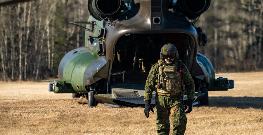 A member of the Canadian Armed Forces dressed in camouflage walks away from a large CH-147 Chinook helicopter with its cargo ramp open.