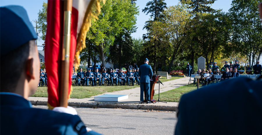 A formal outdoor ceremony with uniformed Canadian Armed Forces members and others seated, and an RCAF speaker is at the podium. 