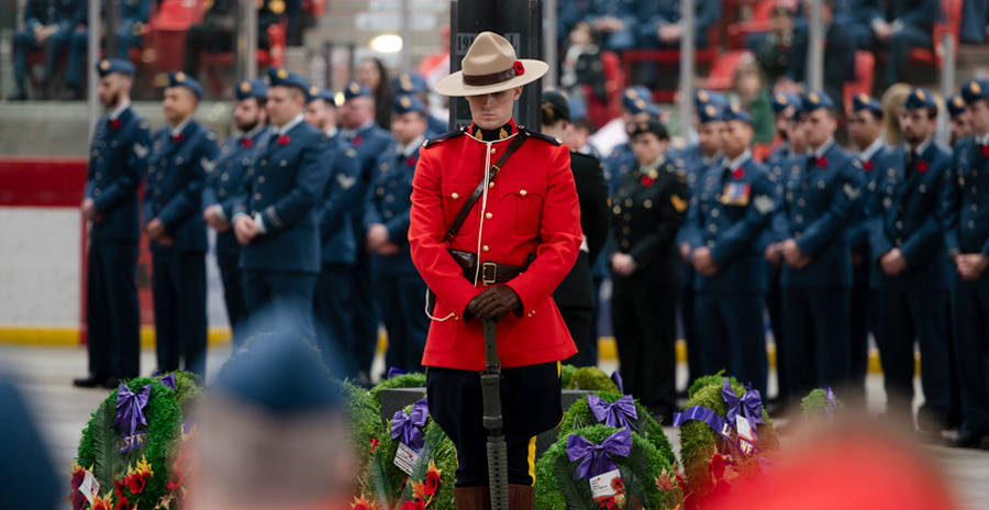 A member of the RCMP stands in front of a cenotaph, with members of the RCAF in the background.