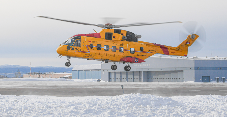 An RCAF helicopter painted in the search and rescue yellow paint scheme flying over a snowy airfield near hangars.