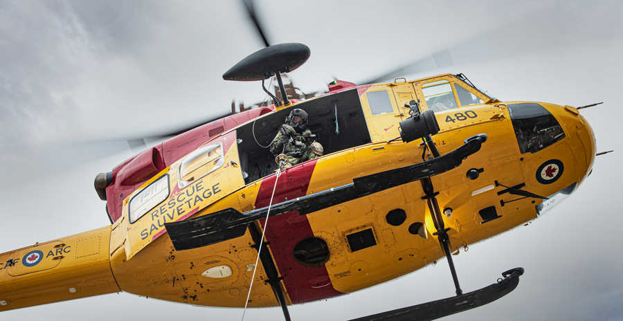 An RCAF search and rescue helicopter in the yellow and red paint scheme in flight with a crew member lowering a cable.