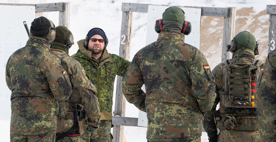 German Army personnel with their backs to the camera take direction from an RCAF member who is speaking at a firearm range.