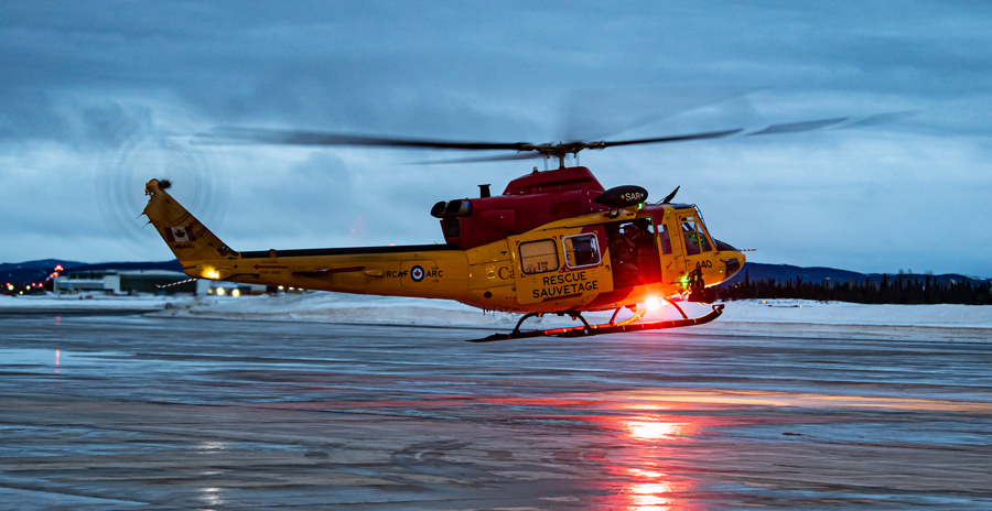 An RCAF red and yellow helicopter hovers over a wet airfield at dusk.