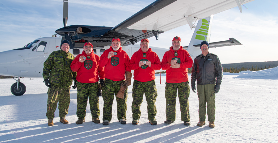 RCAF members pose with Canadian Rangers in front of a Twin Otter aircraft on a snow-covered airfield.