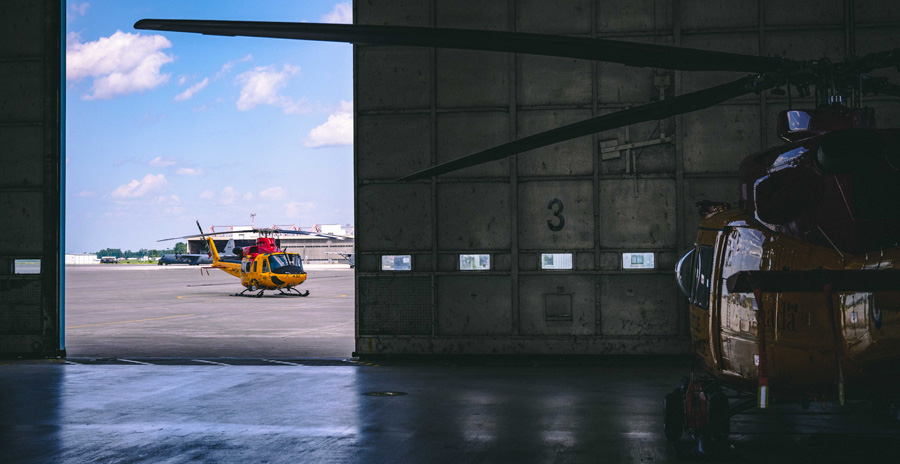 A yellow search and rescue helicopter inside an aircraft hanger and another one is outside the hangar on the tarmac under a blue sky.