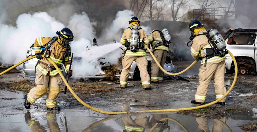 Three firefighters in full protective gear spray water on burning vehicles, while thick smoke rises from the wreckage.