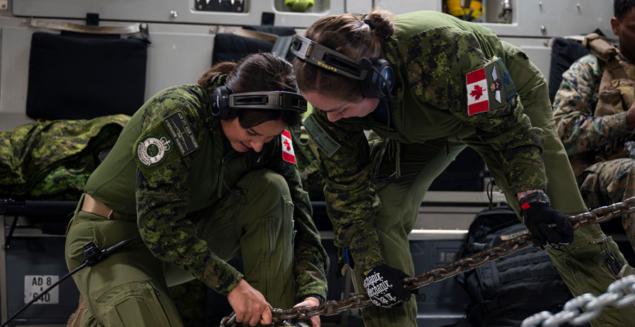 Two military members in uniform secure heavy chains to the floor inside an aircraft.