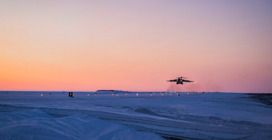 A large military aircraft takes off from a snow-covered runway at sunset.