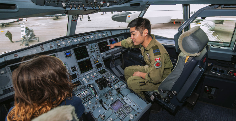 Inside the cockpit of a large aircraft, a military member points to a screen on the instrument panel while another person observes.