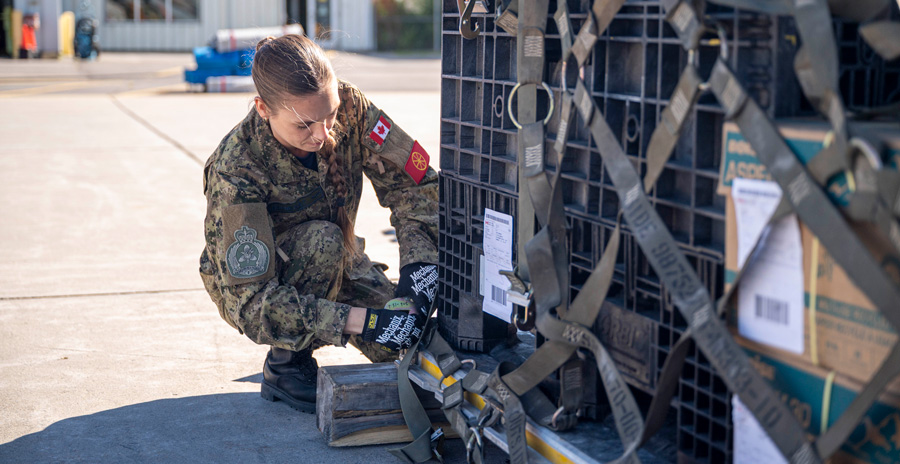 Person in military uniform secures straps on a large cargo pallet outdoors, with a building and equipment visible in the background.