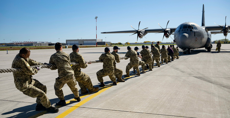 A group of uniformed personnel pull a thick rope attached to a large military transport aircraft on a runway.